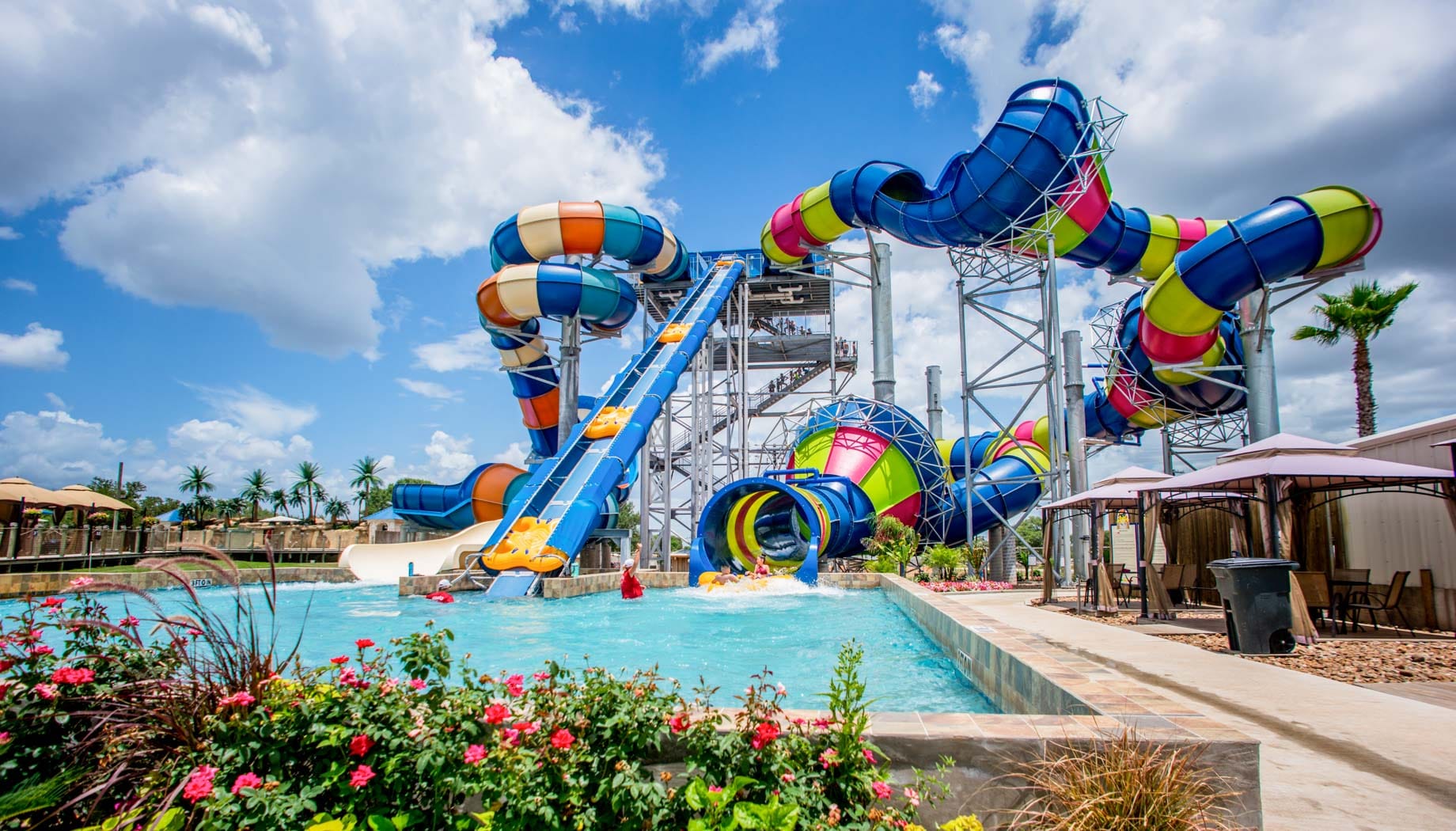 loco-mono-flowers A colorful water park features a large, winding waterslide ending in a clear blue pool surrounded by deck chairs, cabanas, and palm trees under a partly cloudy sky.