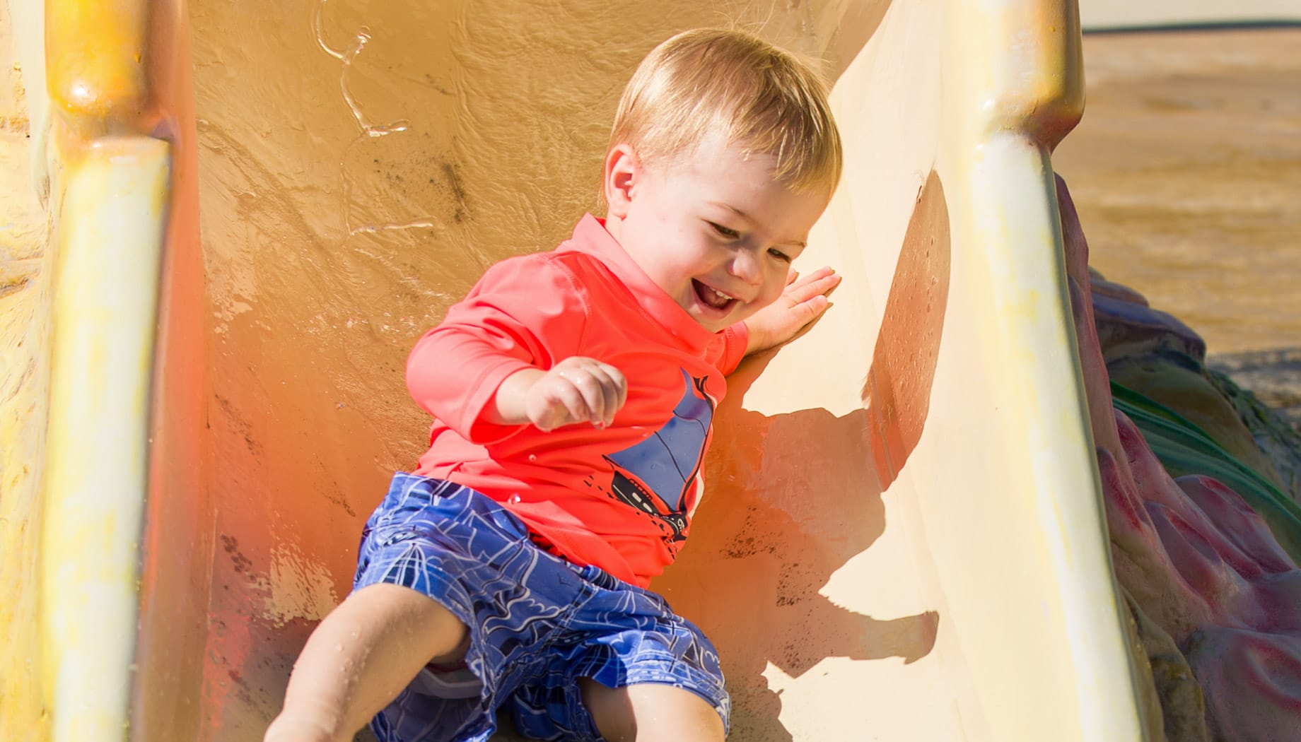 toddler-bay-boy Toddler boy in a red shirt and blue swim trucks sliding down a small water slide.