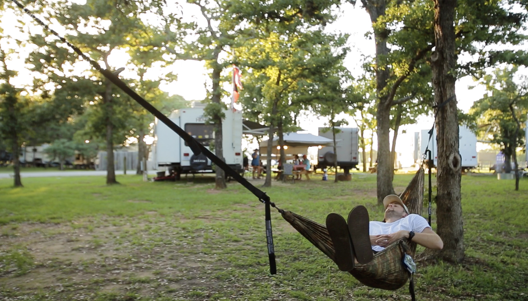 Blog 2 A person relaxes in a hammock tied between two trees in a campsite with parked RVs and people in the background.