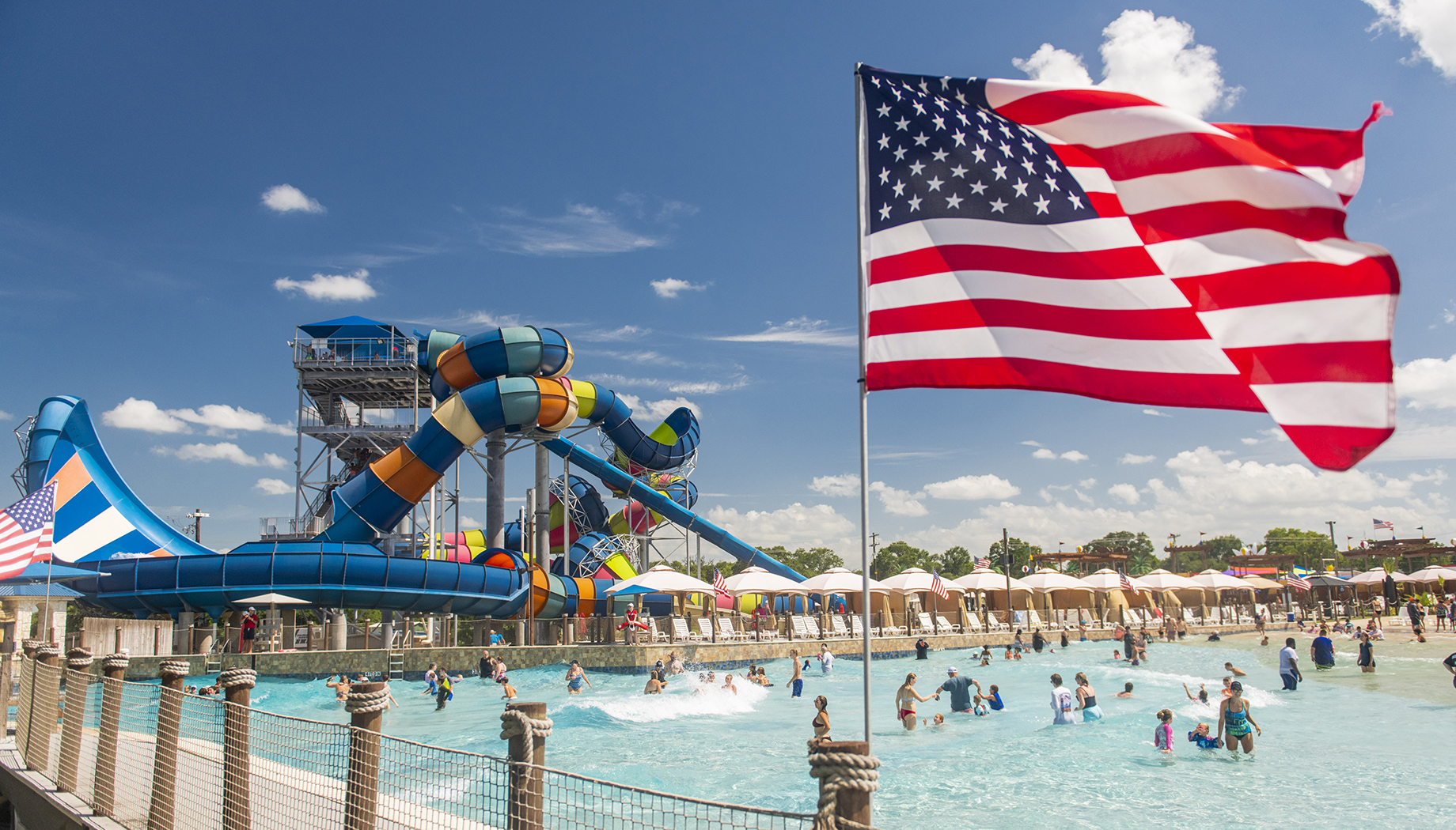 BLOG People enjoying a day at a water park with several slides and a wave pool. An American flag waves in the foreground under a blue sky.