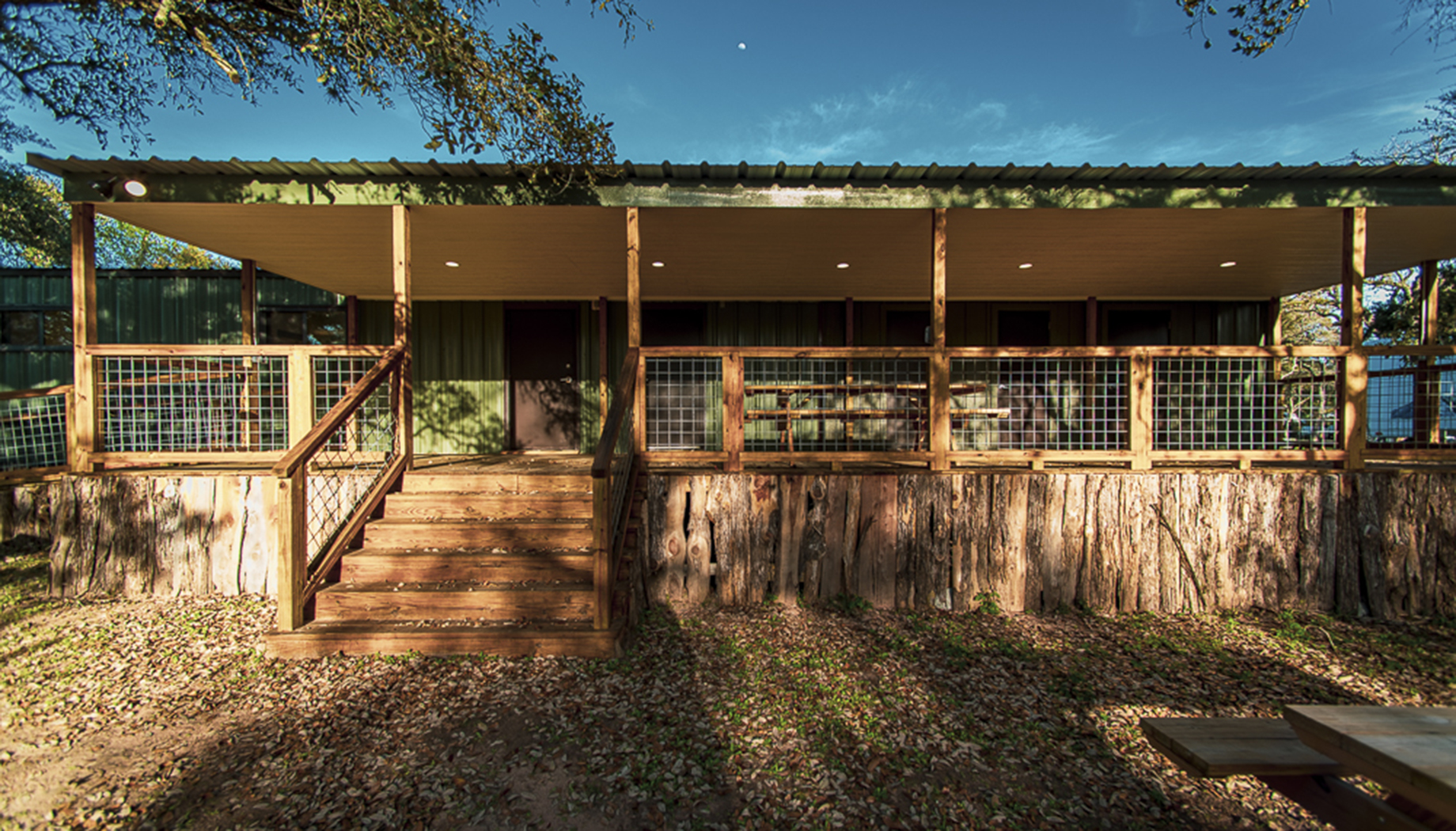 BLOG A green cabin with a metal roof and a wooden porch with stairs leading up to the entrance, surrounded by trees and fallen leaves on the ground.