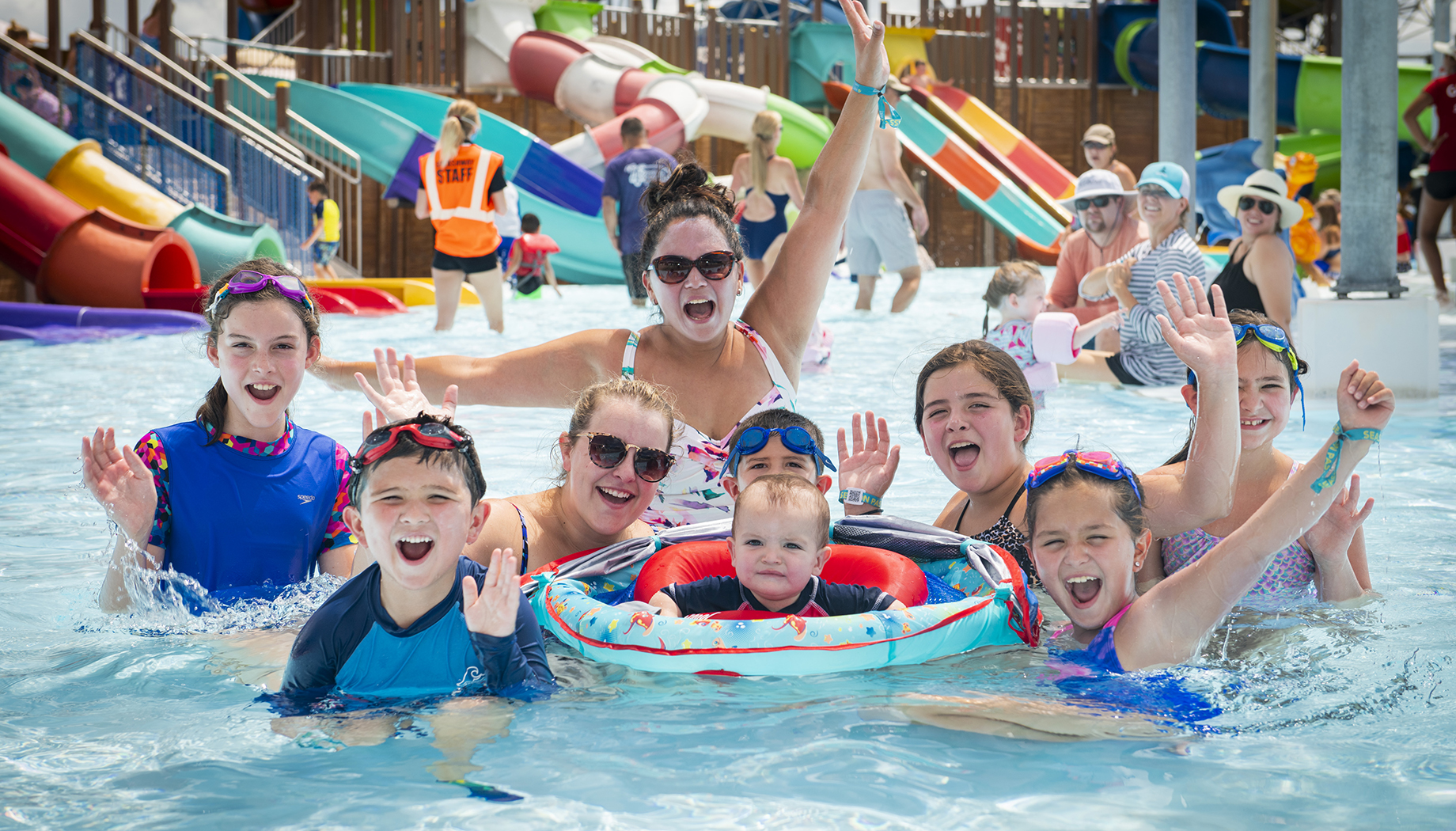 BLOG A group of people, including children and adults, are enjoying themselves in a water park pool. They are all smiling and waving, with colorful water slides visible in the background.