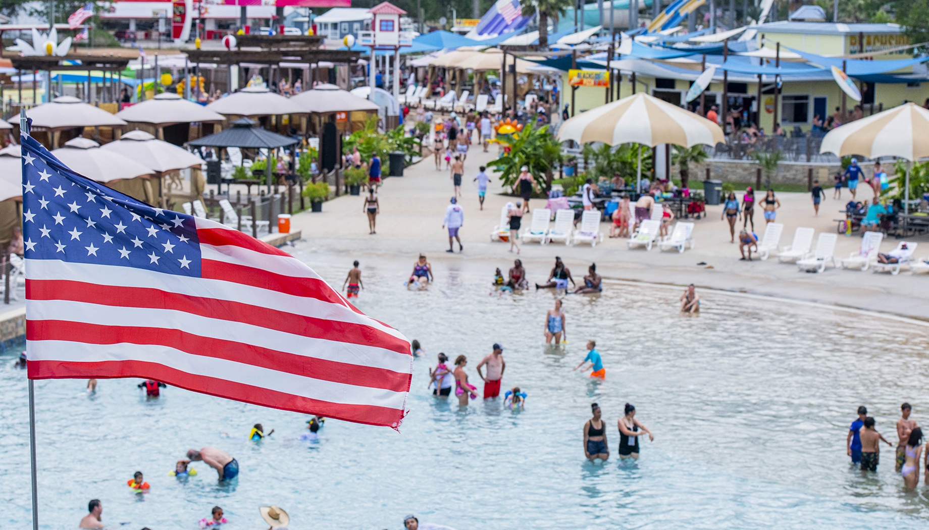 Military and Veteran Savings A large American flag flies over a crowded water park, with people swimming, sunbathing, and walking around. Various water rides and concession stands are visible in the background.