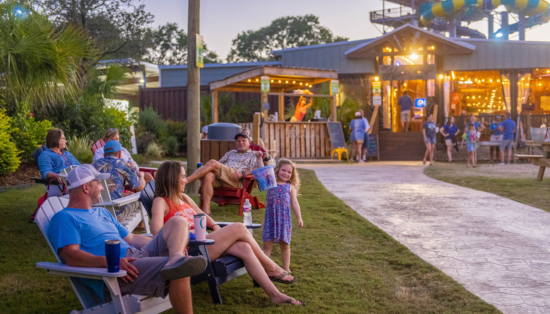 BLOG A group of people relaxes on lawn chairs outside a well-lit building at dusk. A young girl stands smiling in the center, and people can be seen conversing and moving in the background.